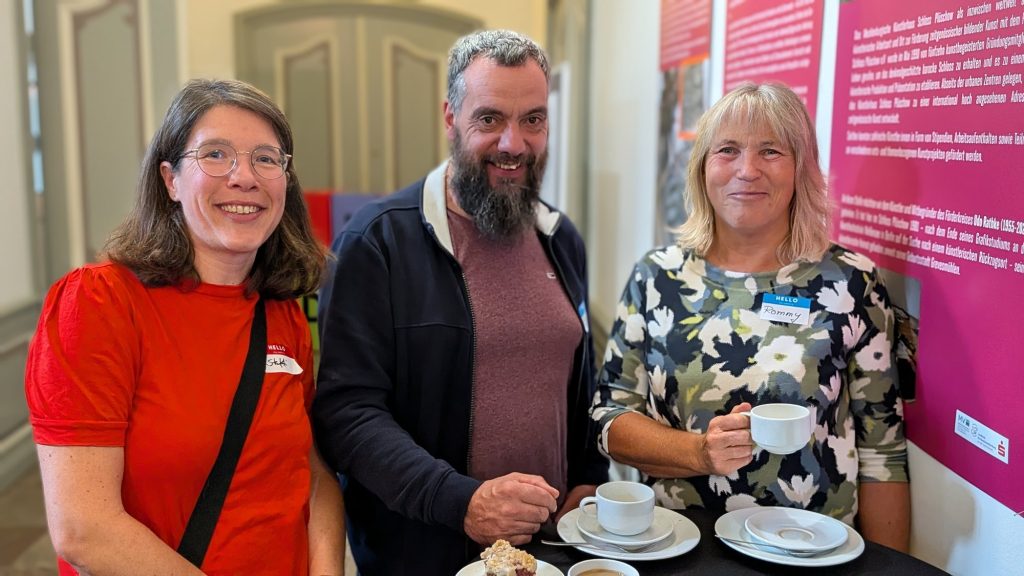 Drei Teilnehmende mit Kaffeetassen vor einer Projektwand
