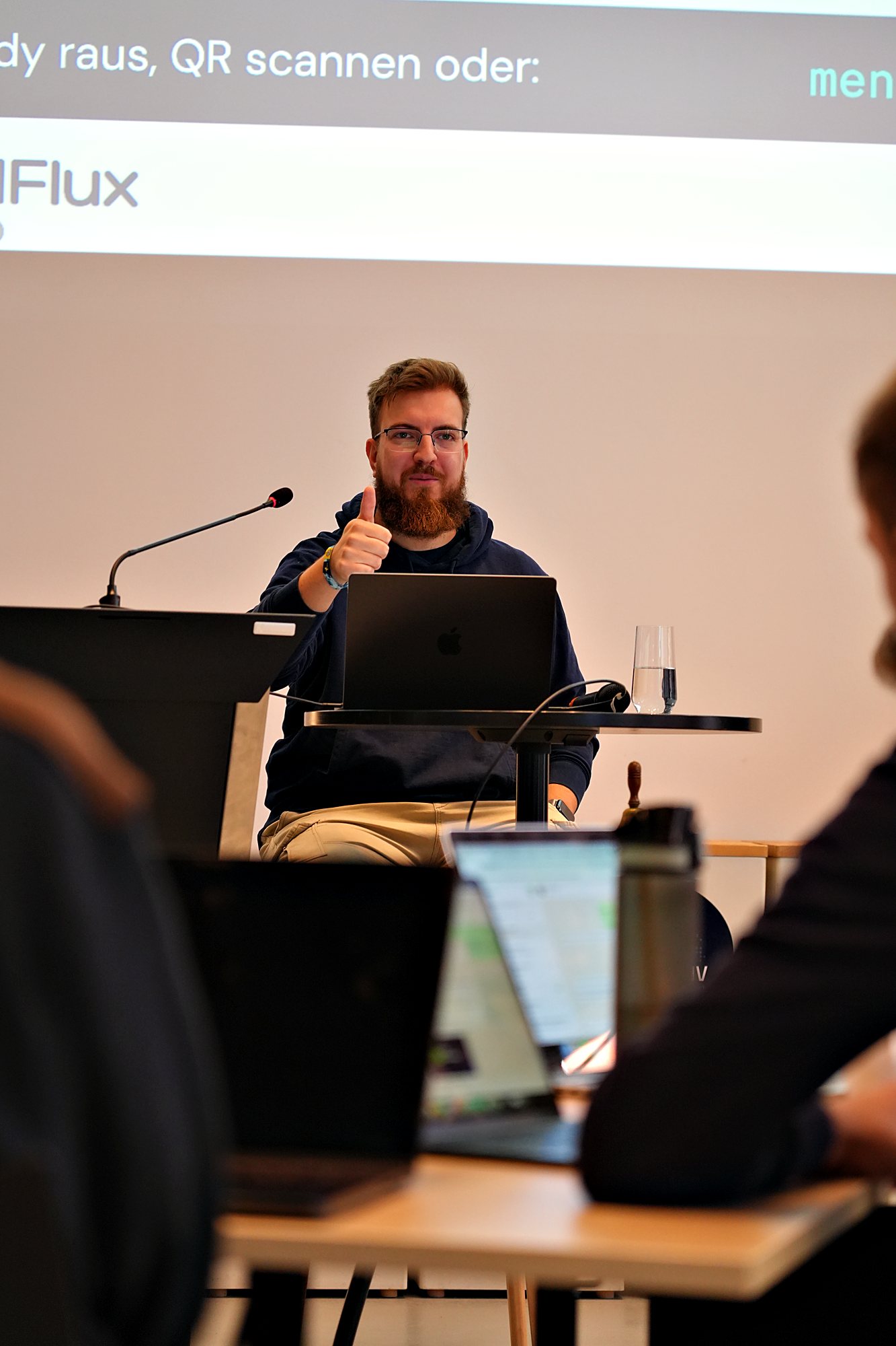 Simon Glowik am Stehtisch hebt den Daumen, im Hintergrund die Leinwand mit dem Menti-Code