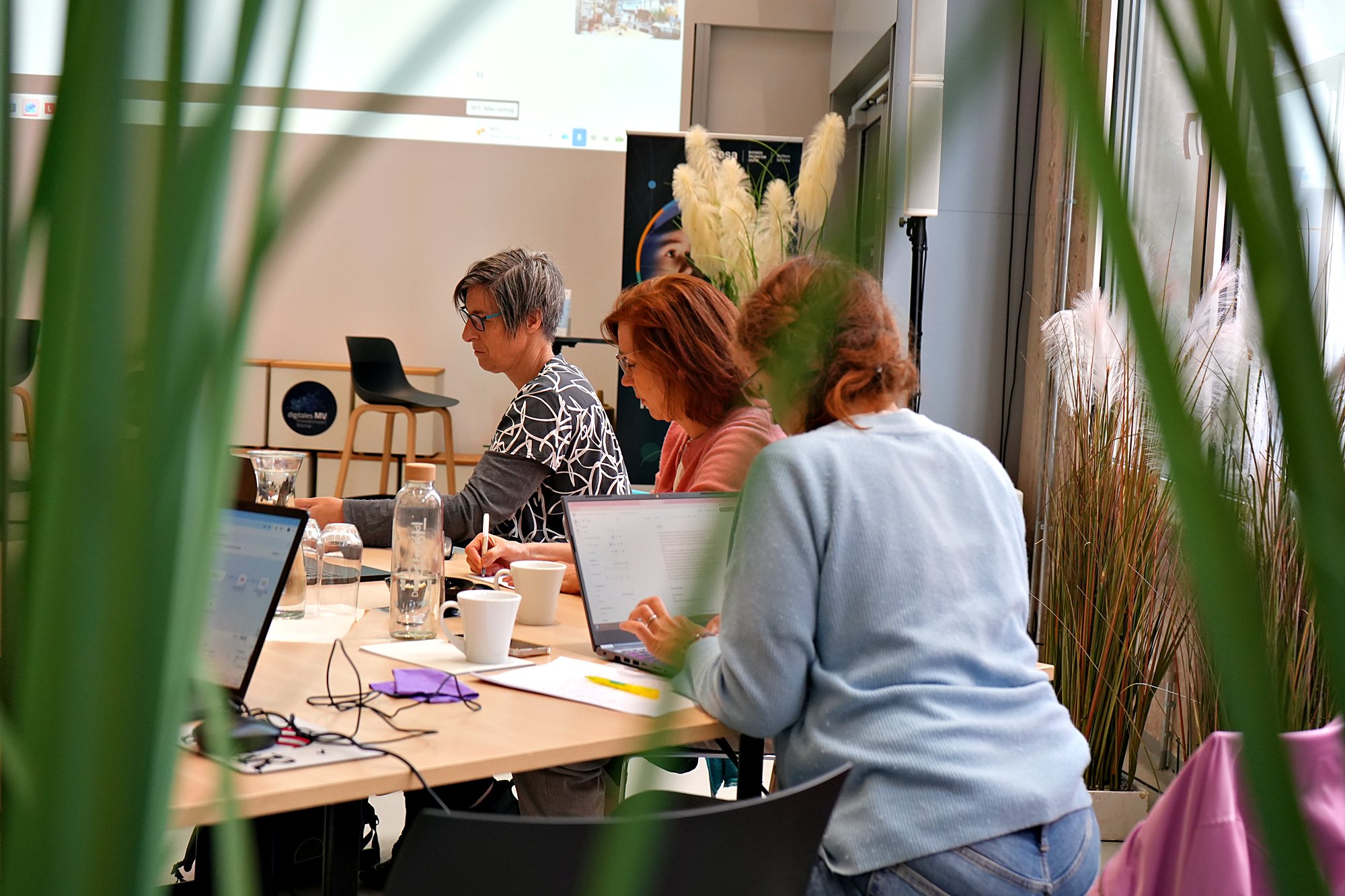 Teilnehmende arbeiten am Tisch, durch Pflanzen im Vordergrund fotografiert