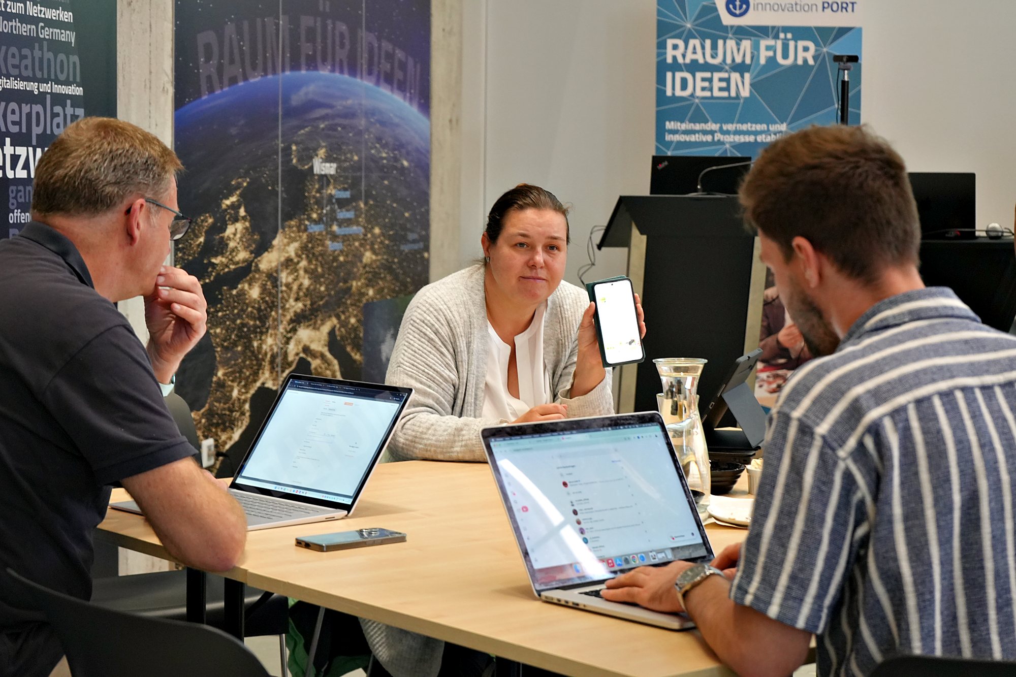 Drei Teilnehmende am Tisch mit Laptops, eine Teilnehmerin zeigt ihr Smartphone