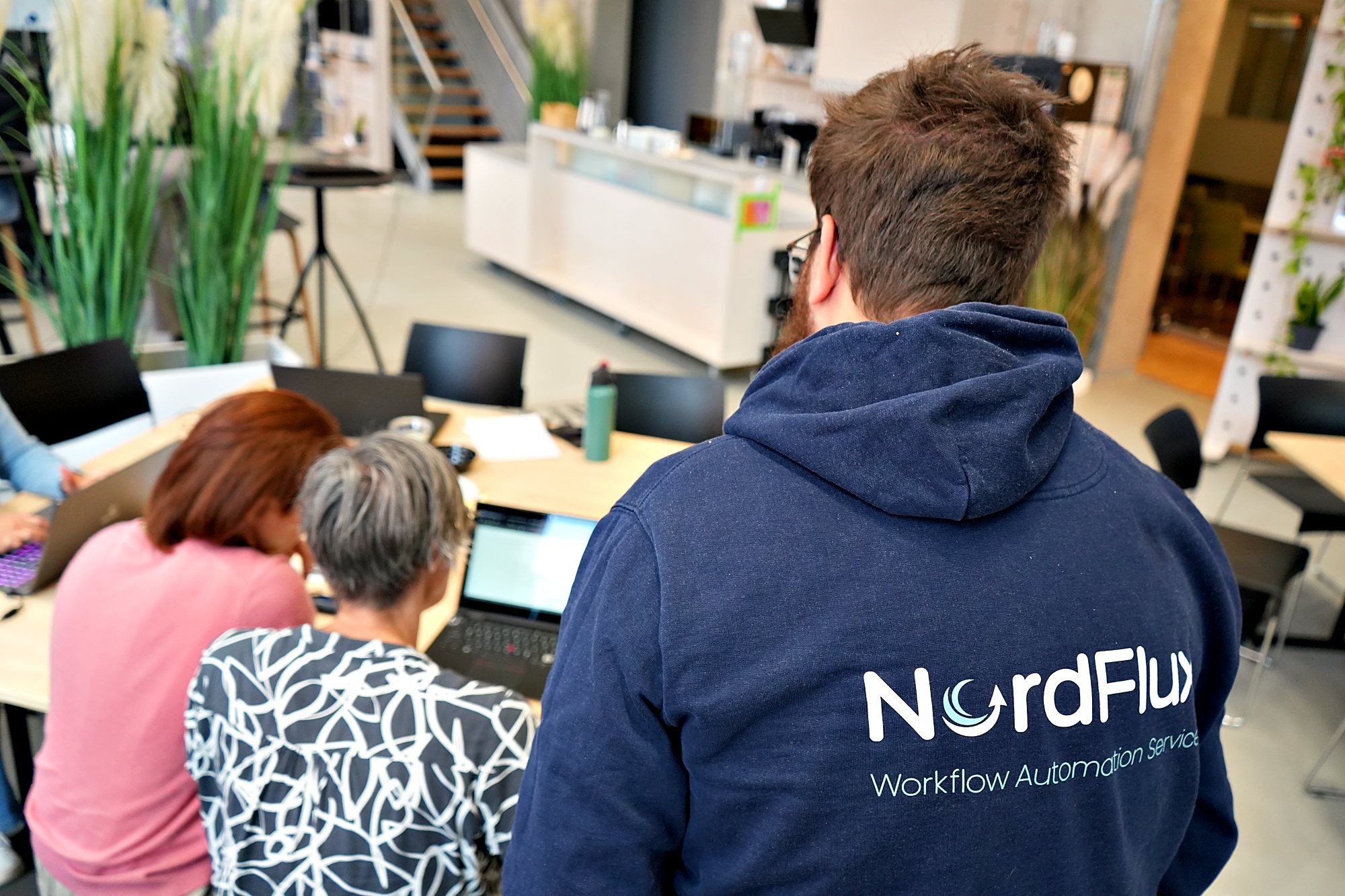 Simon Glowik von hinten im Kapuzenpullover mit NordFlux-Logo, am Tisch arbeiten zwei Teilnehmerinnen