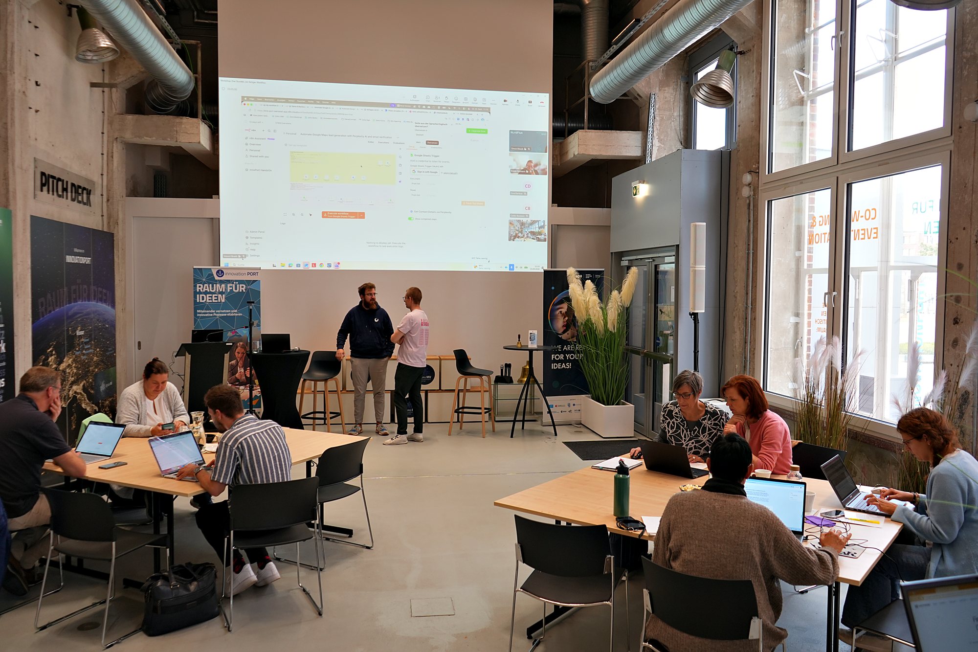 Blick in den Workshopraum, vorn auf der Leinwand ein Workflow in n8n, an den Tischen arbeiten Teilnehmende an Laptops