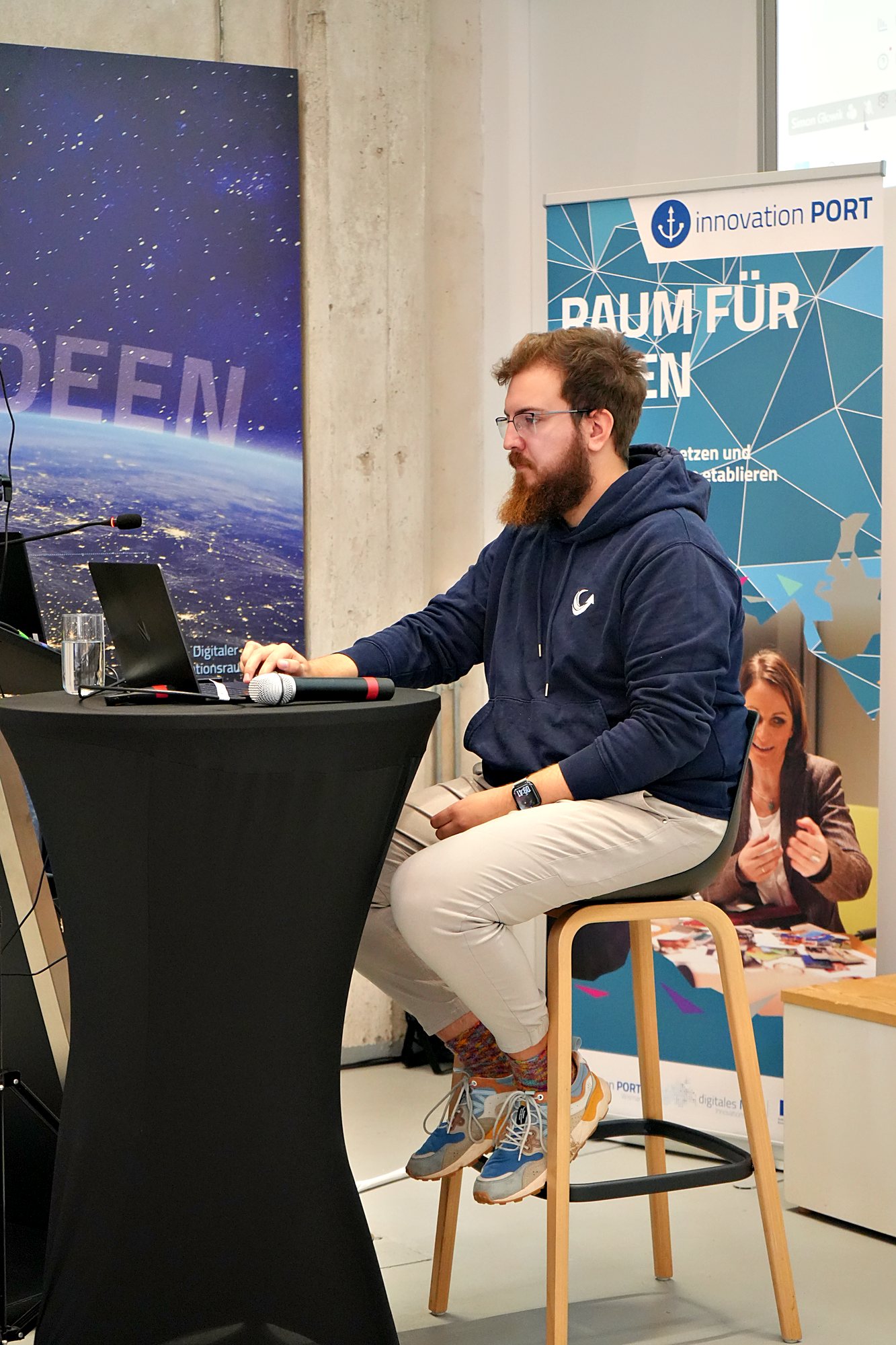 Simon Glowik sitzt am Stehtisch mit Laptop und Mikrofon vor dem Roll-up Raum für Ideen